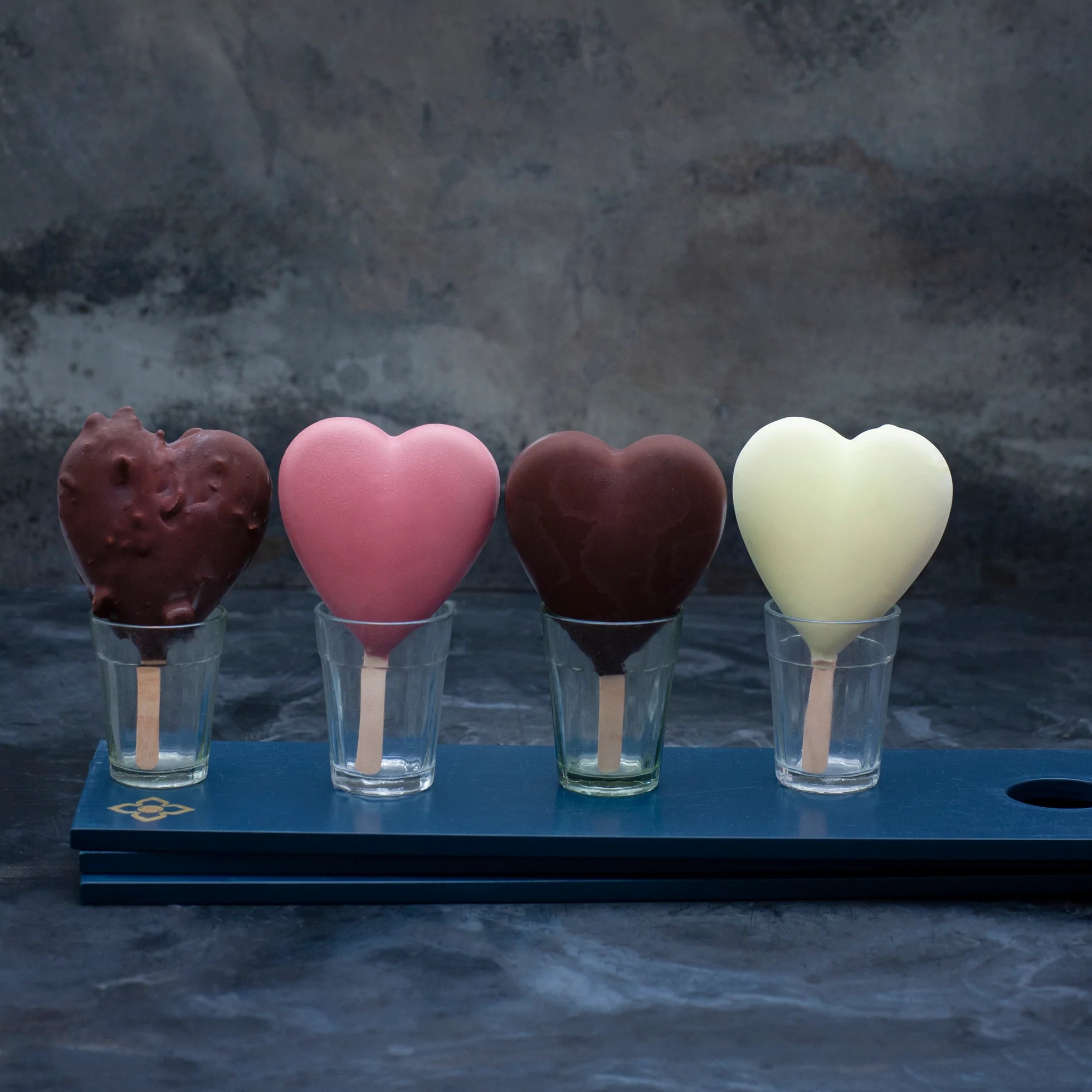 Four heart shaped ice creams on sticks in different colours, standing in little glasses, against a dark background.