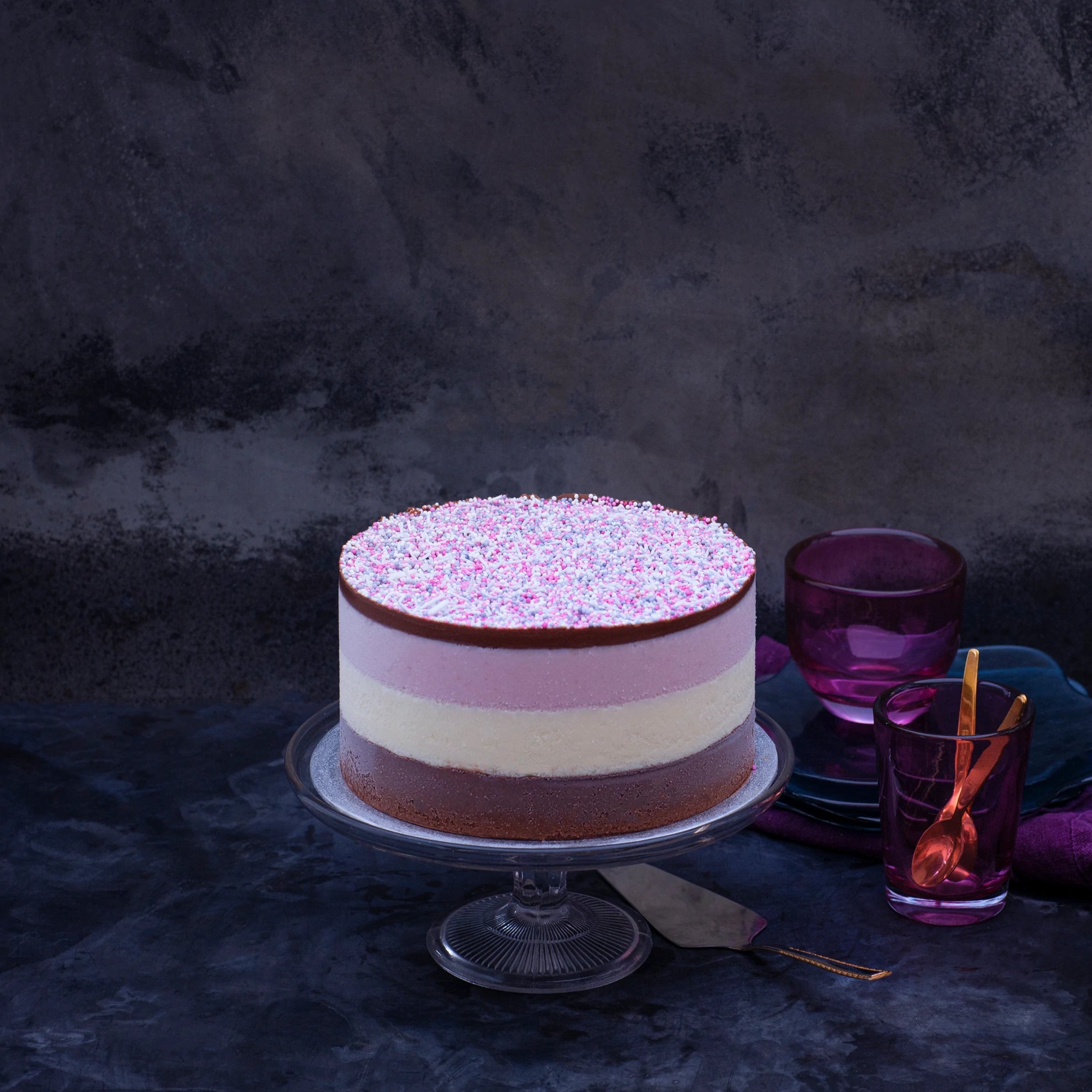 A brown, white and pink layered ice cream cake, topped with sprinkles, set on a glass stand against a dark background.