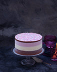 A brown, white and pink layered ice cream cake, topped with sprinkles, set on a glass stand against a dark background.