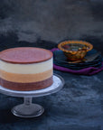 An ice cream cake with white, orange and brown layers, topped with chocolate ganache, on a stand against a dark background.
