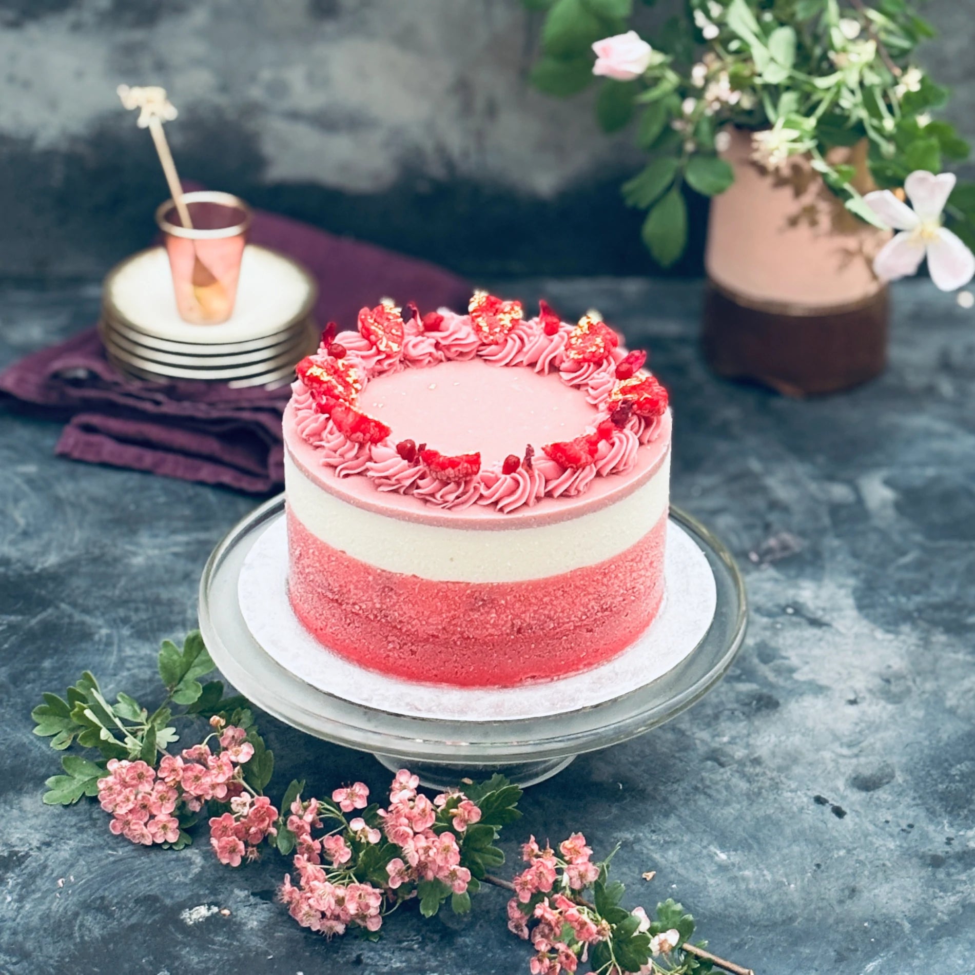 A bespoke ice cream cake with red, pink and white layers, decorated with piped ganache, on a table with pink flowers.