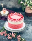 A bespoke ice cream cake with red, pink and white layers, decorated with piped ganache, on a table with pink flowers.