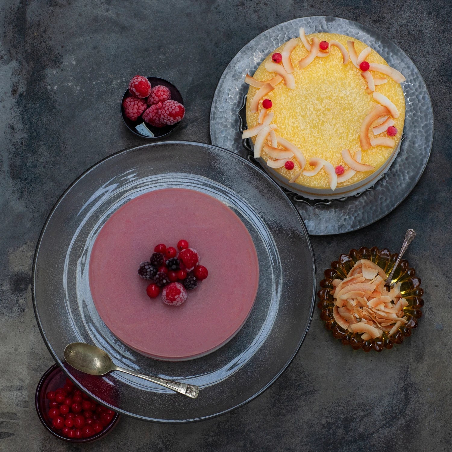 Two sorbet cakes seen from above on a grey background. One is yellow and one is purple, both are decorated with berries.