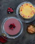 Two sorbet cakes seen from above on a grey background. One is yellow and one is purple, both are decorated with berries.