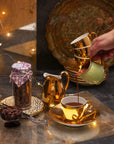 Hot chocolate being poured in a golden cup, next to a glass jar full of chocolate callets