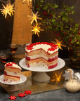 A tall Xmas cake with different coloured layers, topped with white ganache and red flowers, on a festive background.