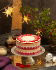 A tall Xmas cake with different coloured layers, topped with white ganache and red flowers, on a festive background.
