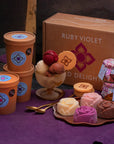 Ice cream scoops in a glass bowl, ice cream roses on a plate, jars of toppings, ice cream tubs and a cardboard box on a purple surface.