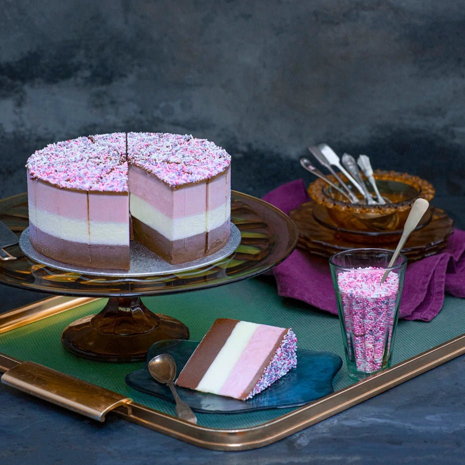 A brown, white and pink layered ice cream cake, topped with sprinkles, set on a glass stand with a slice cut out on a tray.