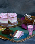 A brown, white and pink layered ice cream cake, topped with sprinkles, set on a glass stand with a slice cut out on a tray.