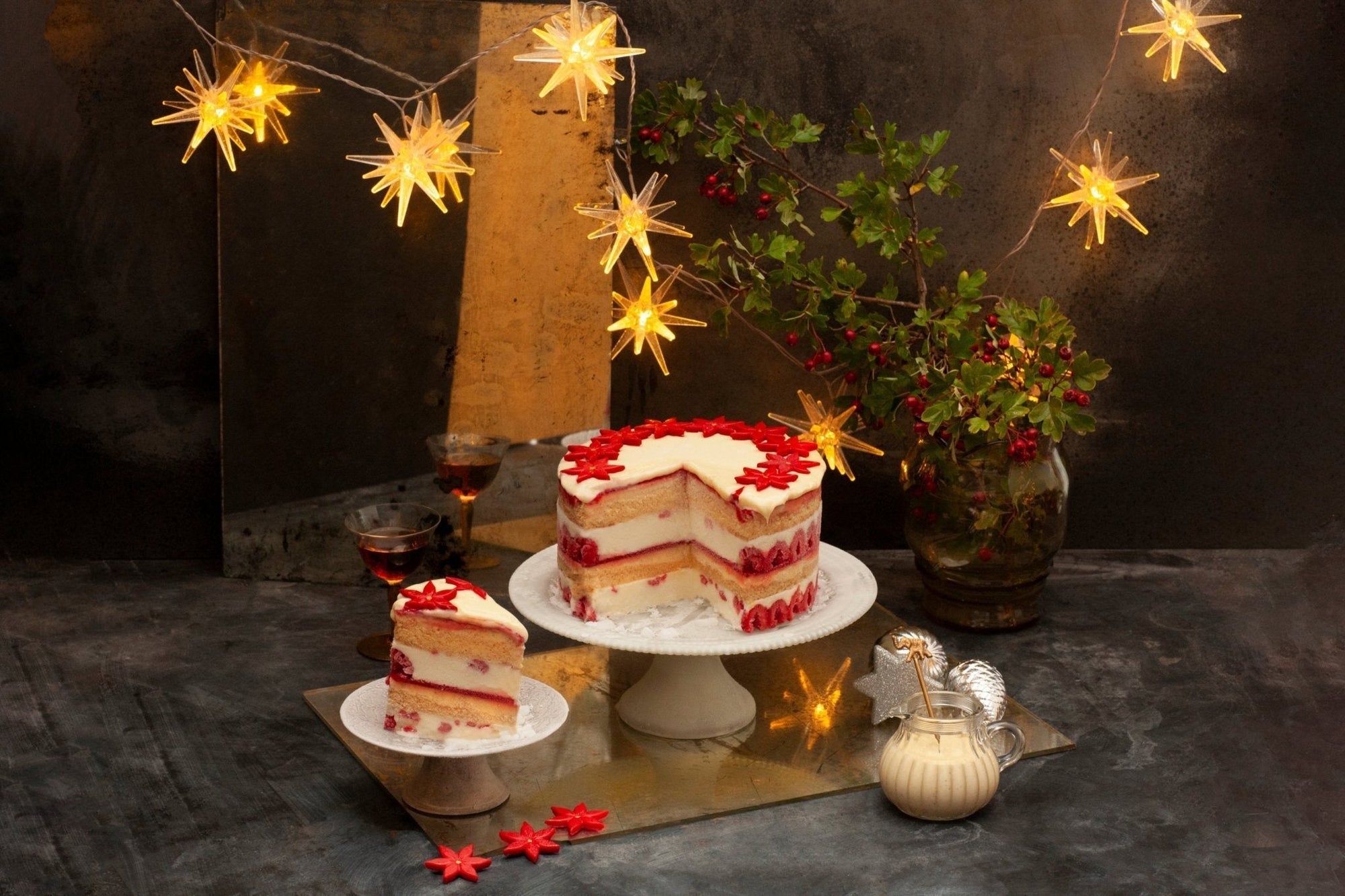 A tall ice cream cake in red and white, with sponge layers, topped with white ganache and red Christmas flower. There is a slice cut out on a plate and a festive background. 