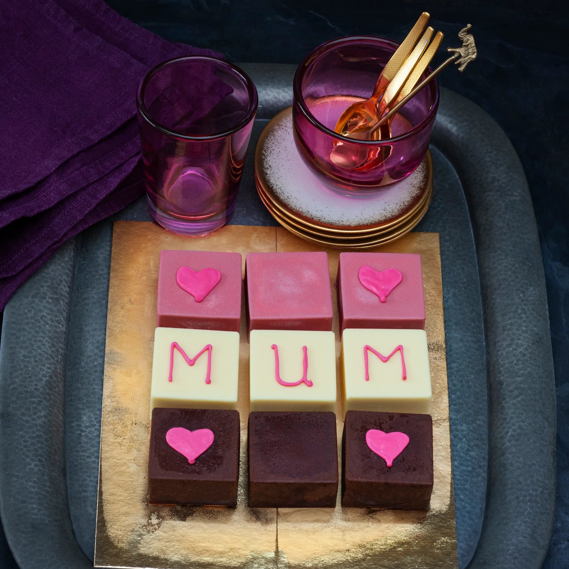 Mini square ice cakes on a tray, 3 covered in ruby chocolate, 3 in white chocolate and 3 in dark chocolate, decorated with hearts and with the word "mum"