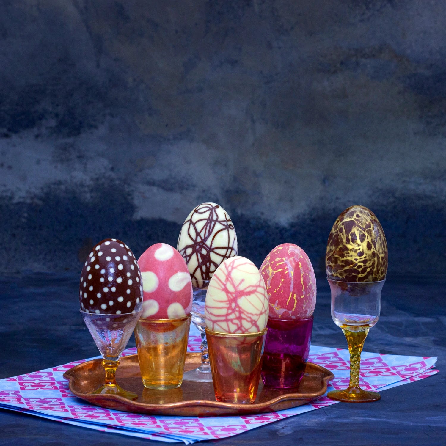 Six easter eggs filled with ice cream and decorated with different motifs sitting in coloured glass cups.