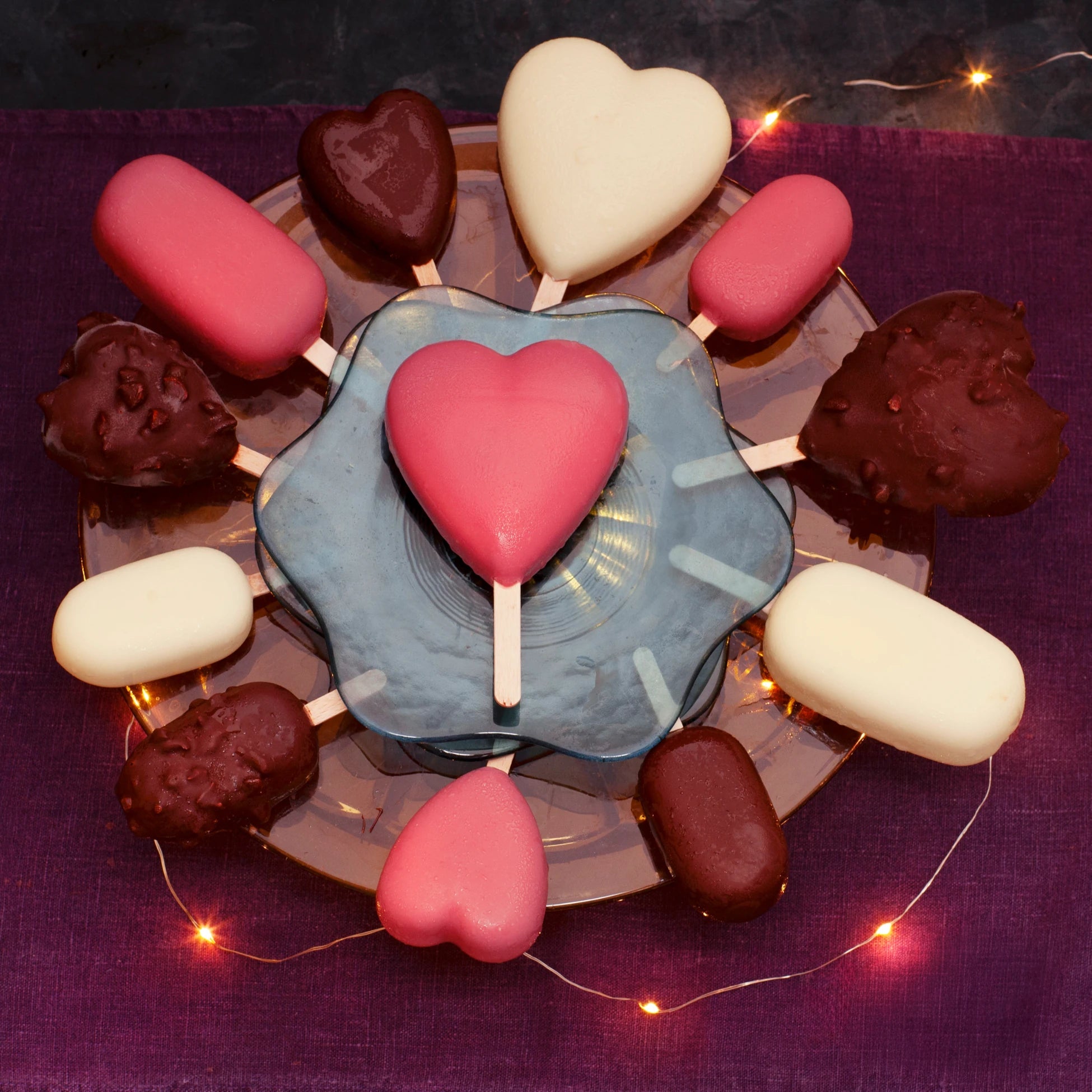 Heart-shaped and regular ice creams on sticks on a plate with a purple background and string lights.