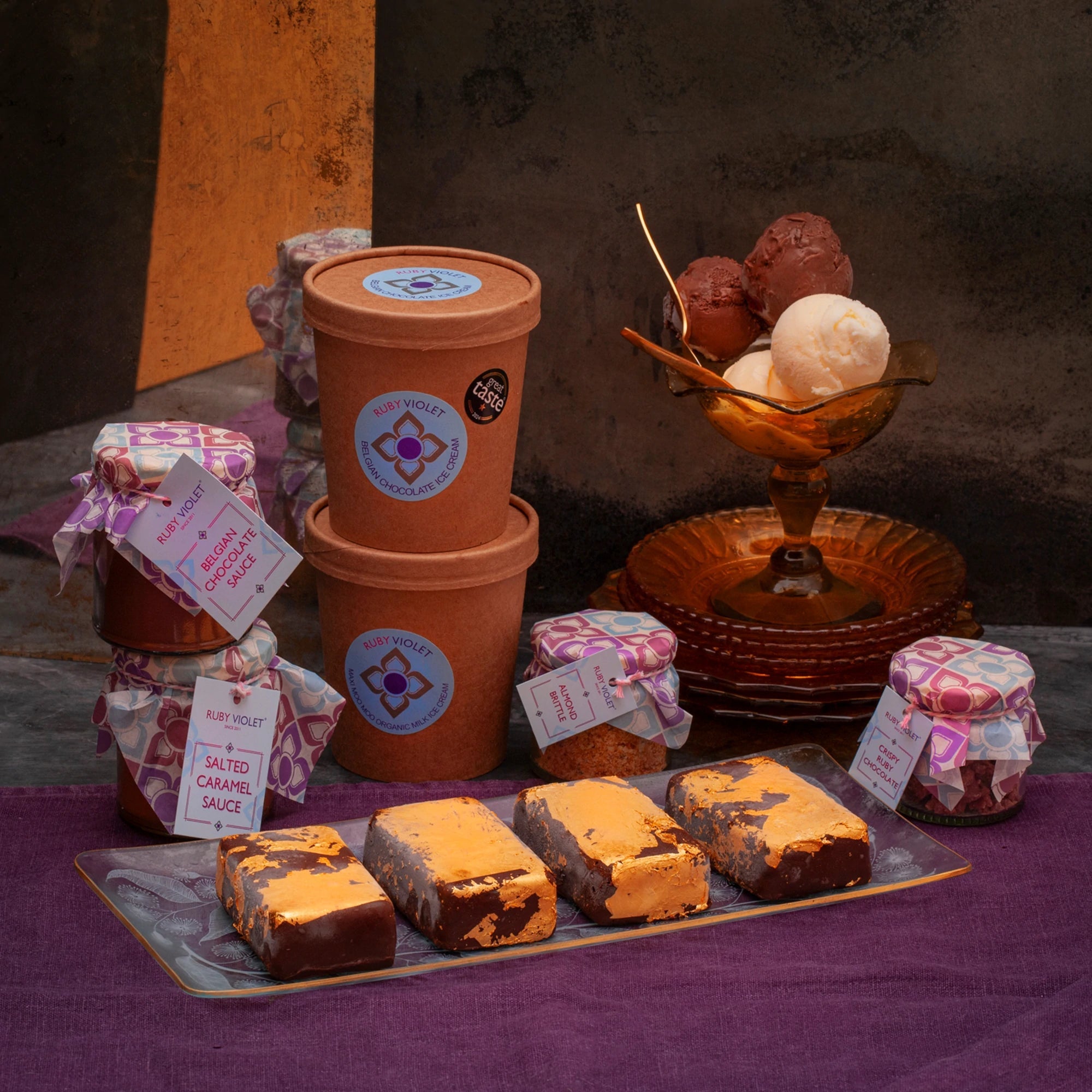 Ice cream tubs, scoops of ice cream, jars of sauce and 4 golden ice cream bullion bars on a purple surface. 