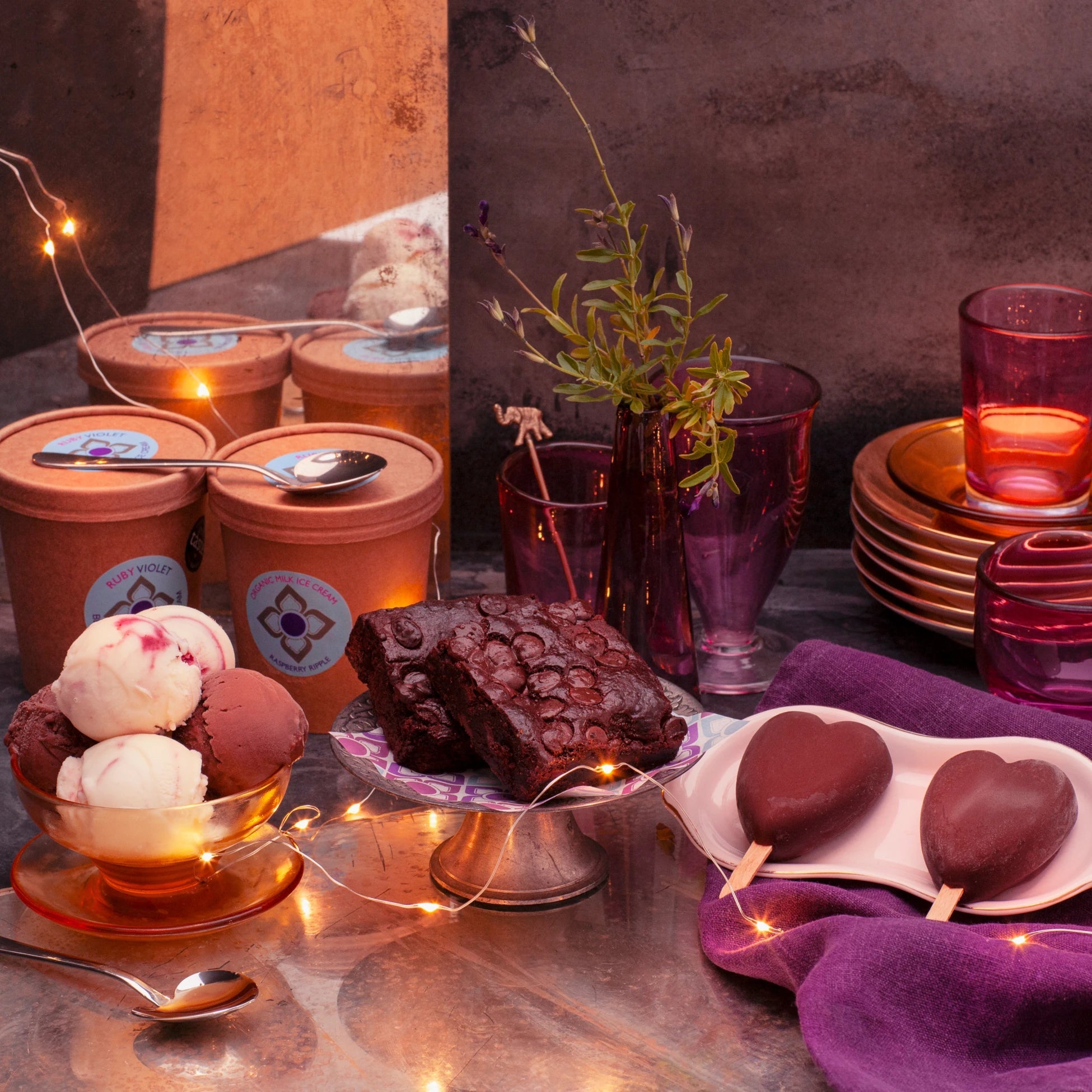 Dessert table with ice cream, brownies, and heart shaped ice cream on sticks on a metallic surface with string lights.