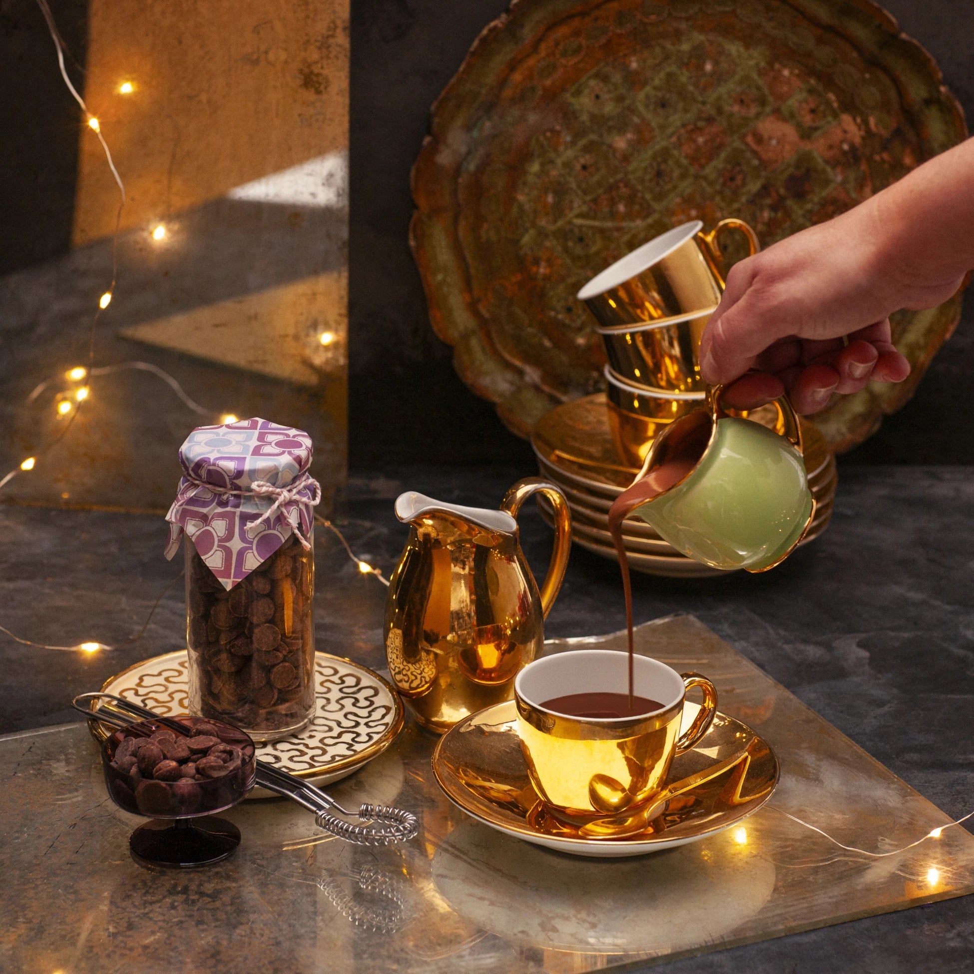 Hot chocolate being poured in a golden cup, next to a glass jar full of chocolate callets