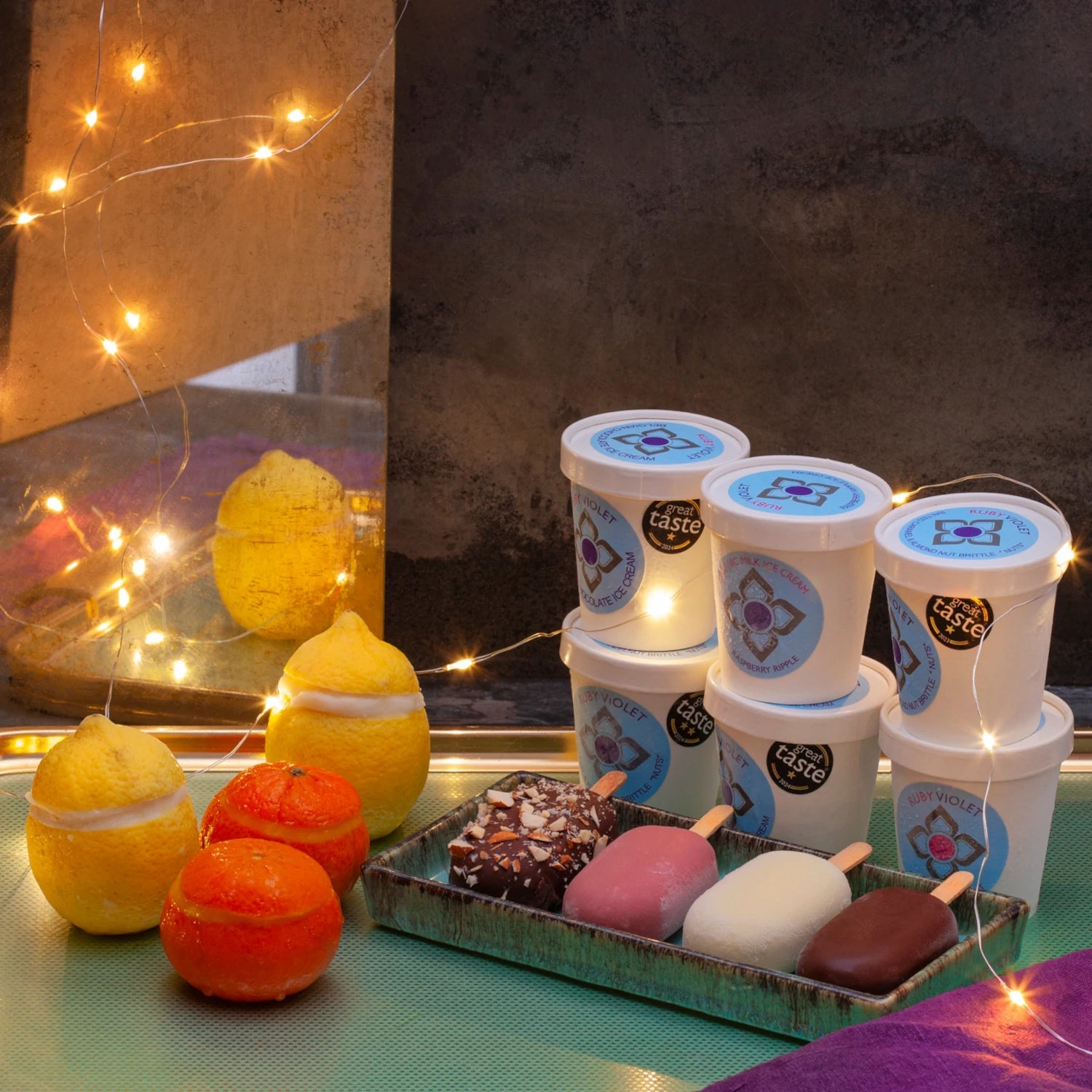 Frozen lemons and clementines, 6 little ice cream tubs piled on each other and 4 colourful ice creams on stick surrounded by fairy lights. 
