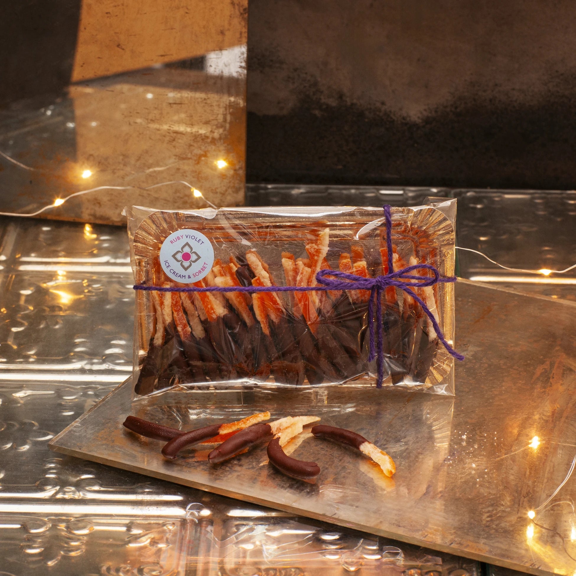 Packaged candied orange peel dipped in chocolate on a gold tray, tied with a purple ribbon ,on a reflective surface with string lights.