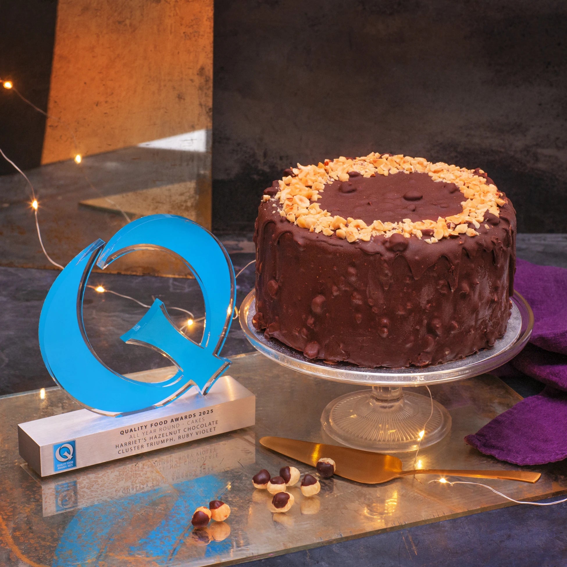 A tall cake coated in chocolate and hazelnut on a glass stand next to a gold Quality Food award