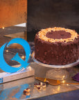 A tall cake coated in chocolate and hazelnut on a glass stand next to a gold Quality Food award