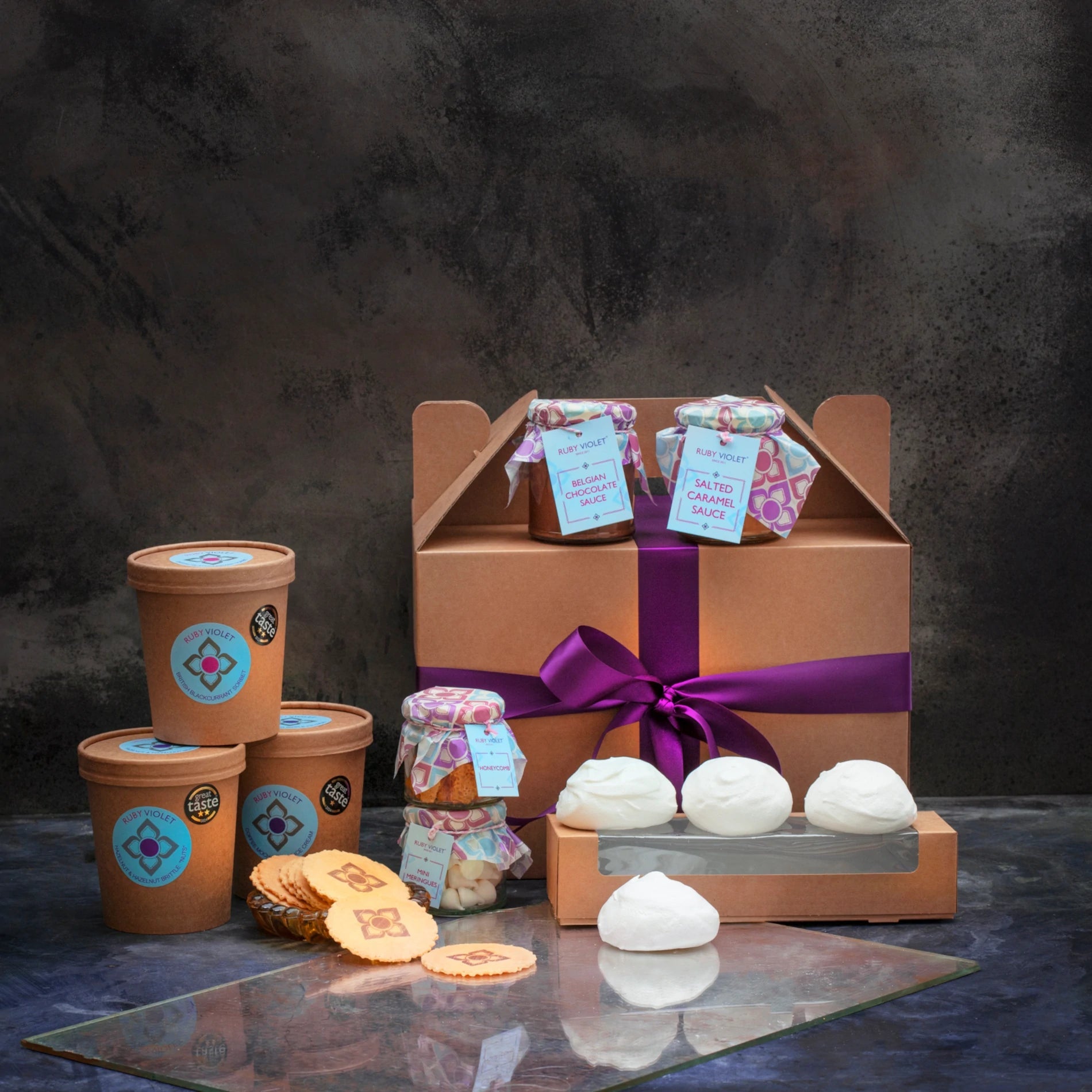 A hamper with 3 ice cream tubs, jars of topping ad sauces, 3 meringues and a gift box against a dark background. 