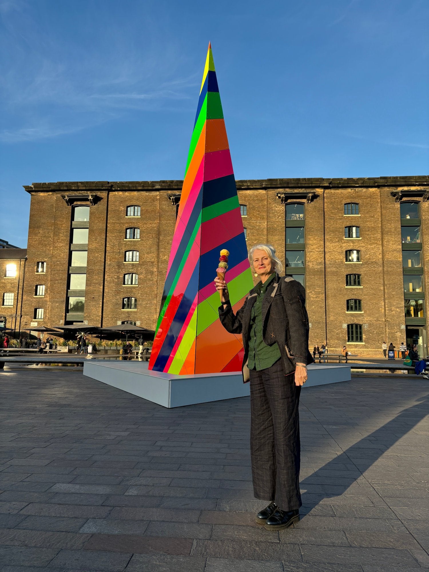 A tall woman standing in front of a colourful structure in London at sunset, holding an ice cream cone