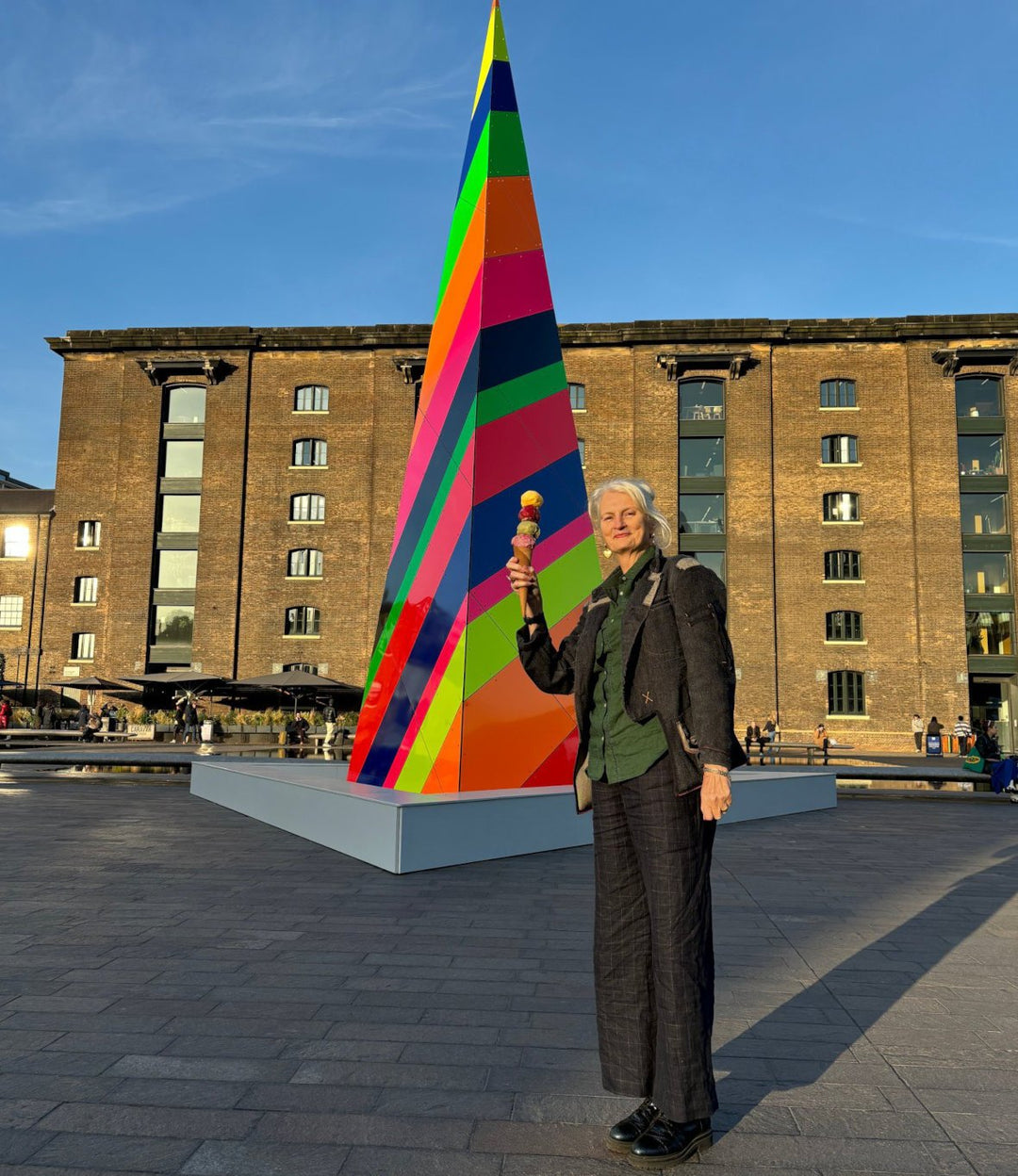 A tall woman standing in front of a colourful structure in London at sunset, holding an ice cream cone