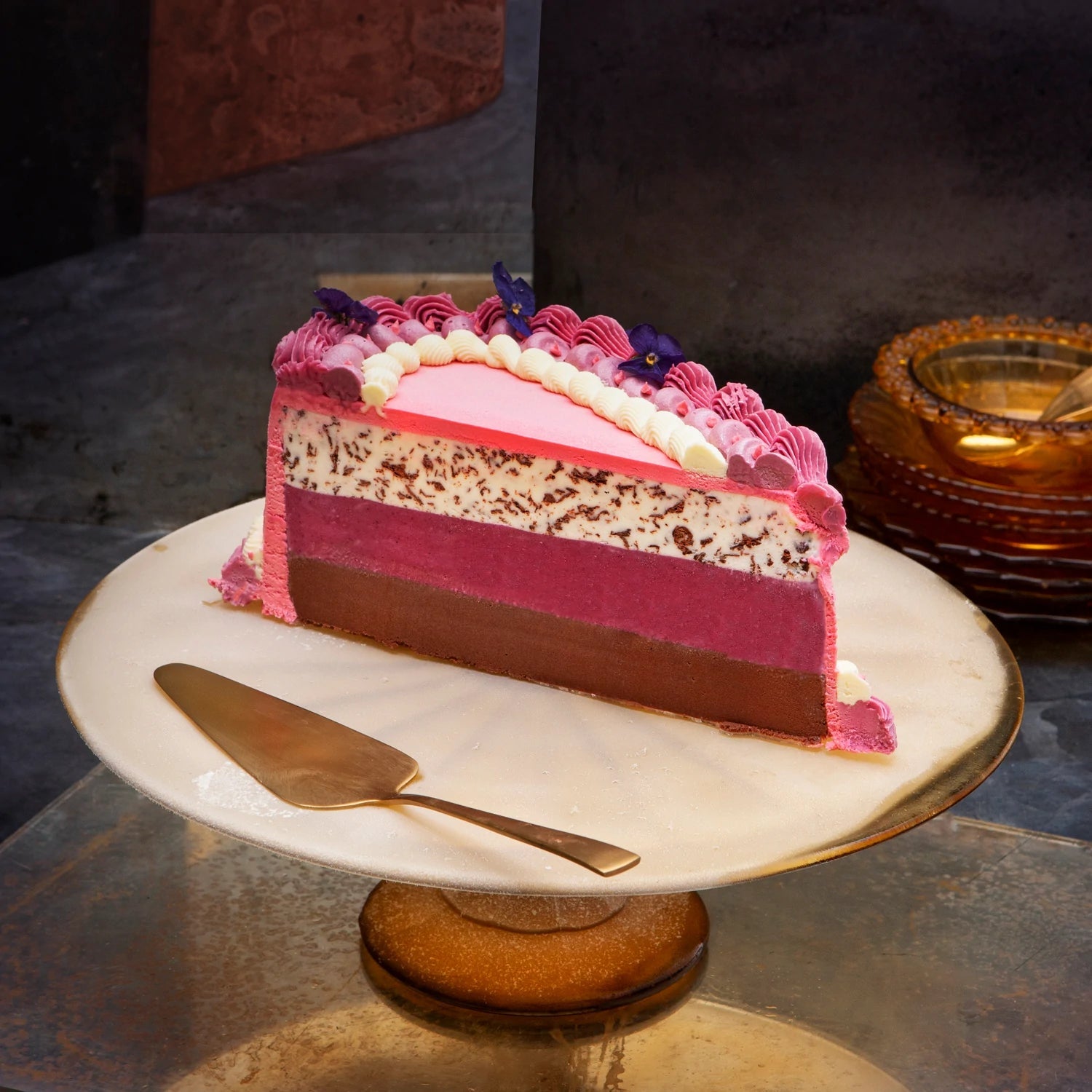 A pink heart shaped cake cut in half to reveal 3 different layers, decorated with piped ganache and violas, on a white stand.