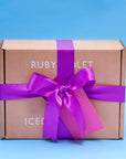 A Ruby Violet cardbord box closed with a purple satin ribbon and matching card seen from the side