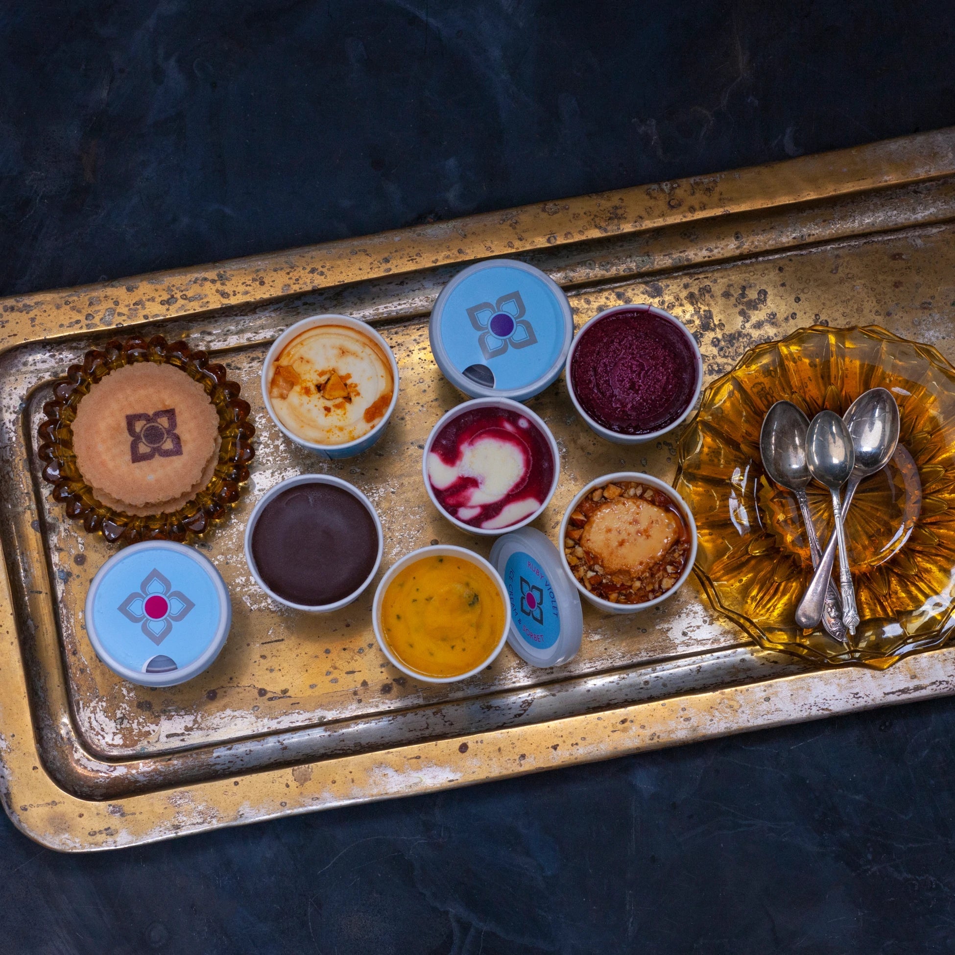 Vintage tray with small tubs of different coloured ice creams and sorbets and a plate with spoons on a dark background
