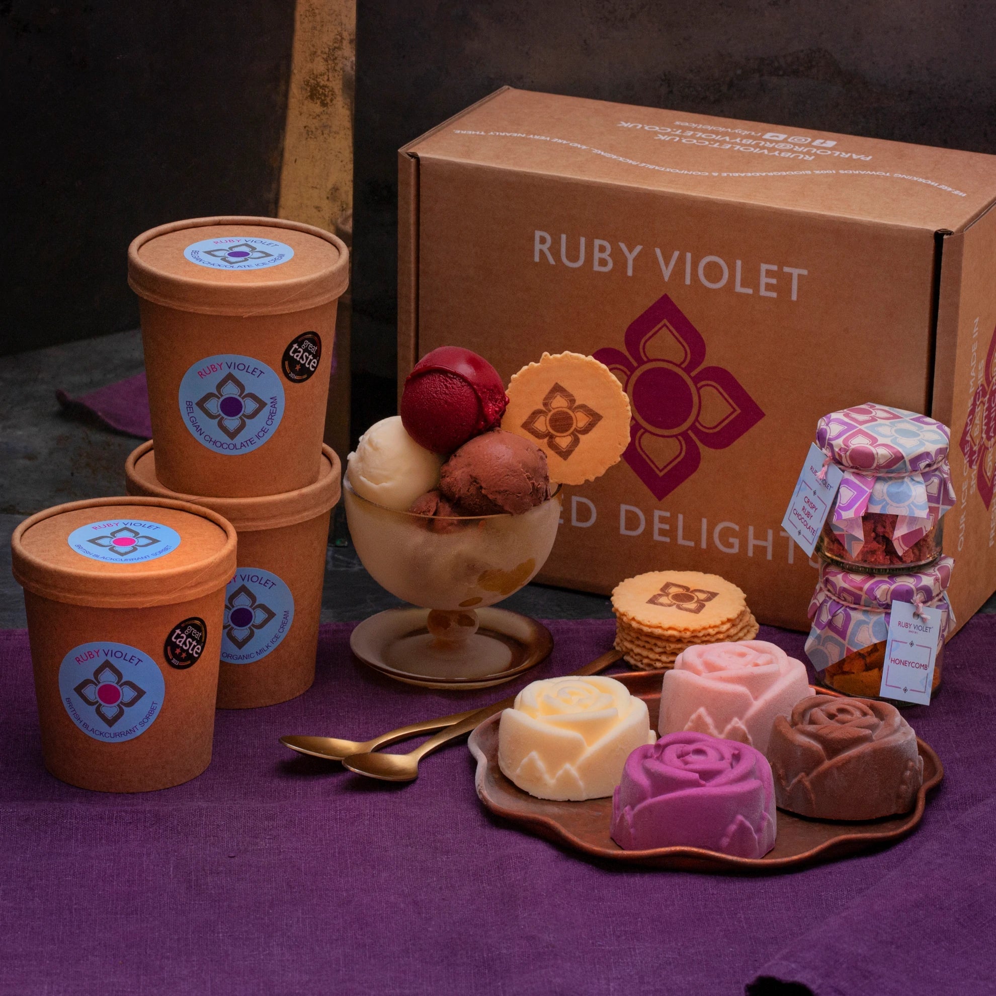 Ice cream scoops in a glass bowl, ice cream roses on a plate, jars of toppings, ice cream tubs and a cardboard box on a purple surface. 
