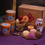 Ice cream scoops in a glass bowl, ice cream roses on a plate, jars of toppings, ice cream tubs and a cardboard box on a purple surface. 