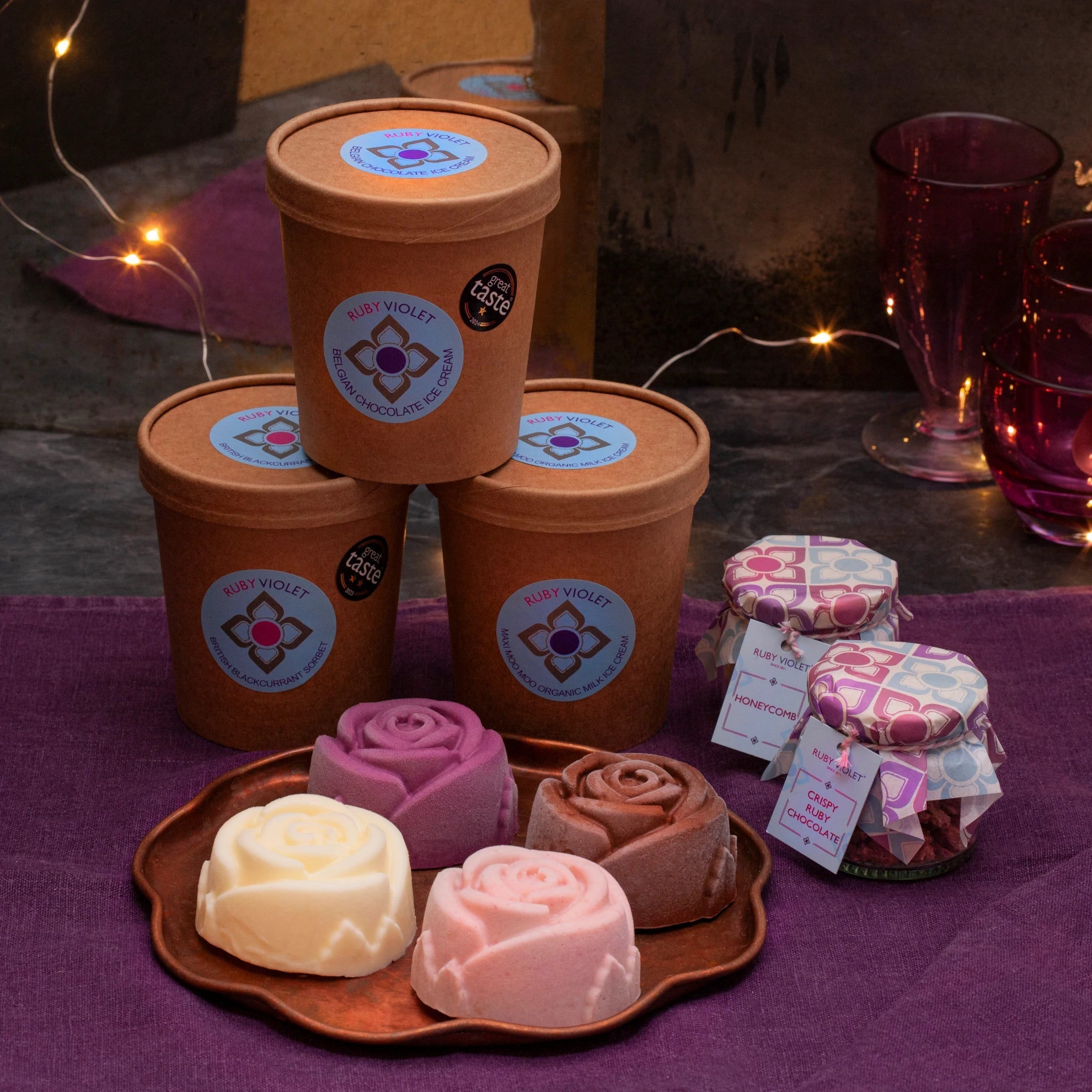 Colourful ice cream roses on a plate, jars of toppings and 3 ice cream tubs on a purple surface. 