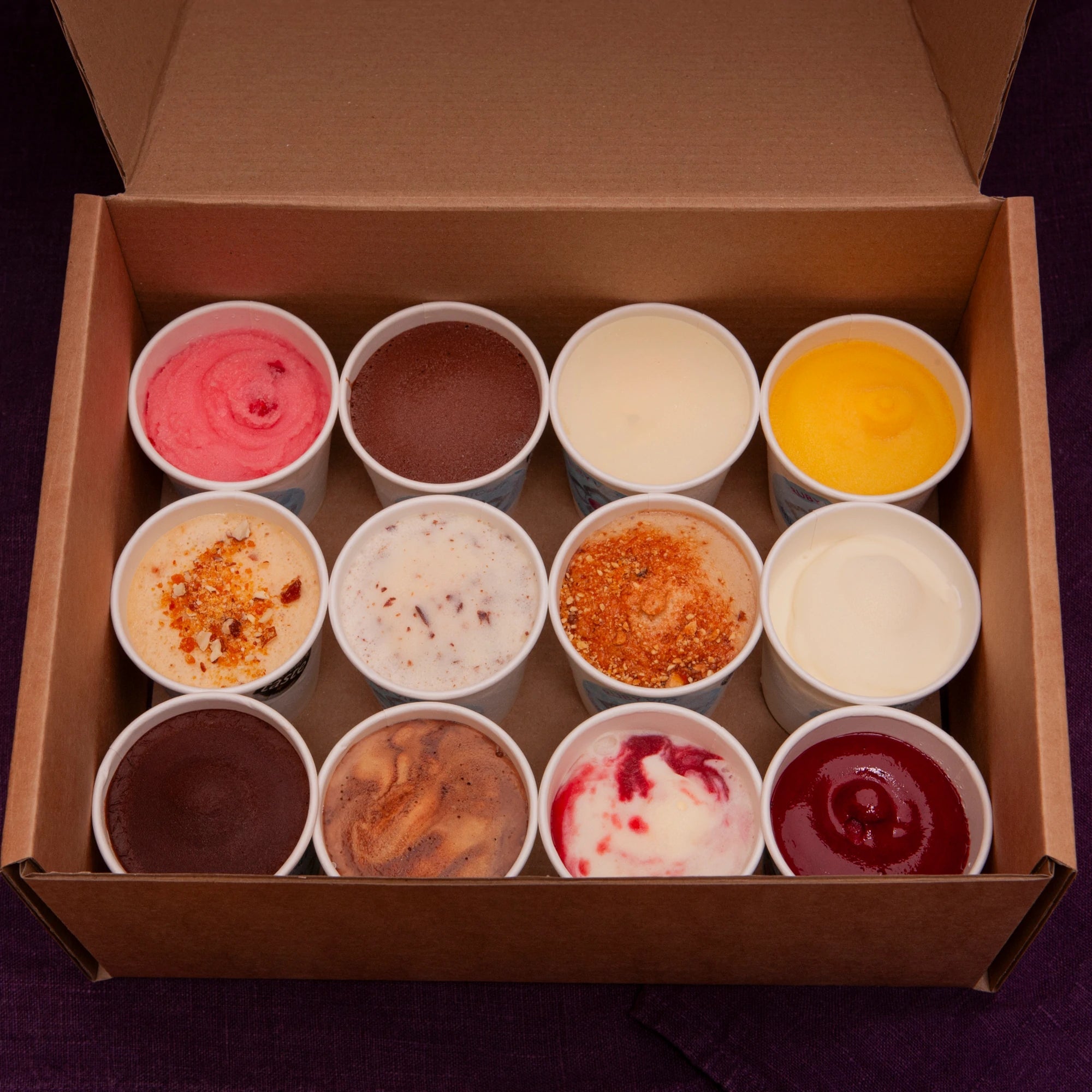 Assorted ice cream flavors in small tubs inside a cardboard box on a dark background