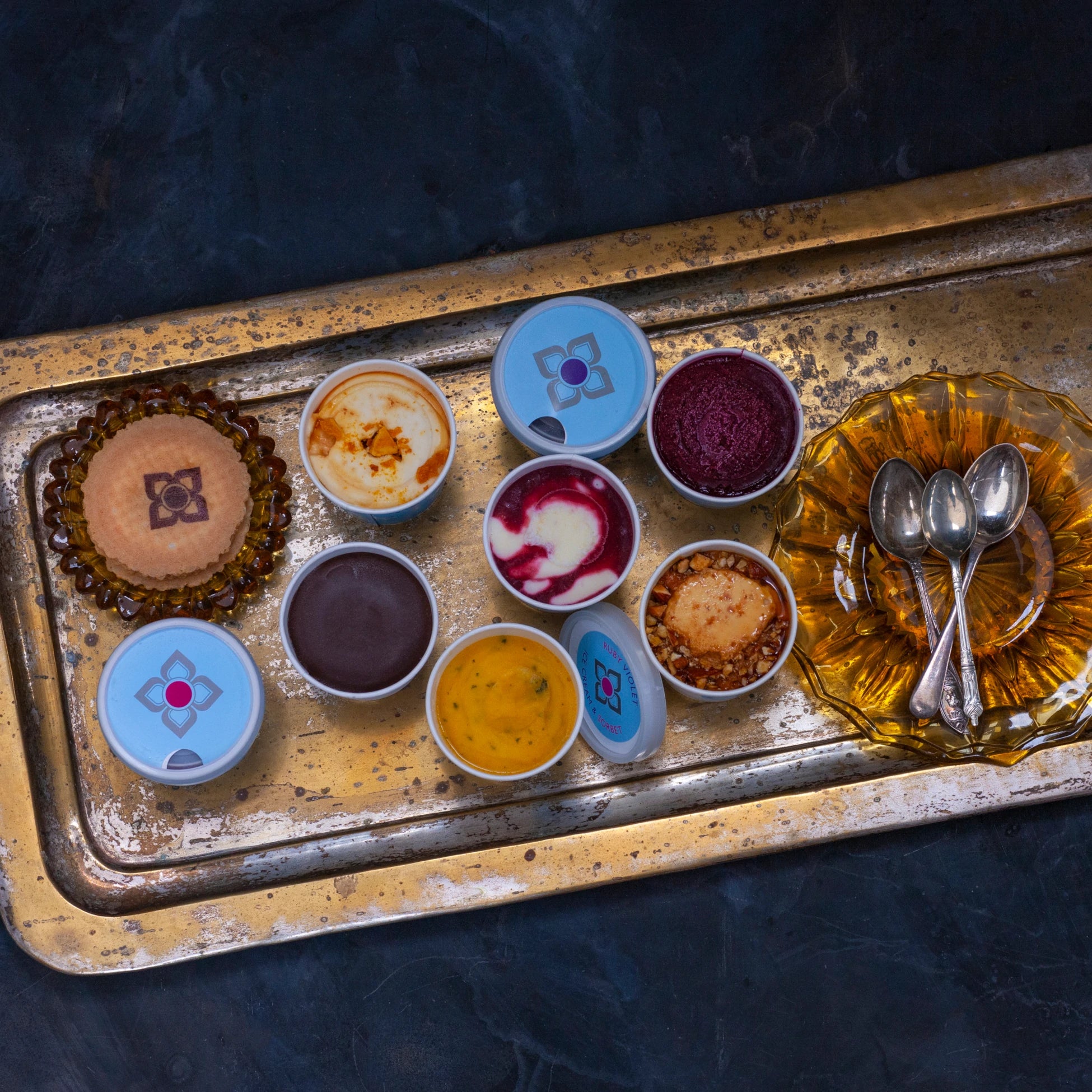 Vintage tray with small tubs of different coloured ice creams and sorbets and a plate with spoons on a dark background
